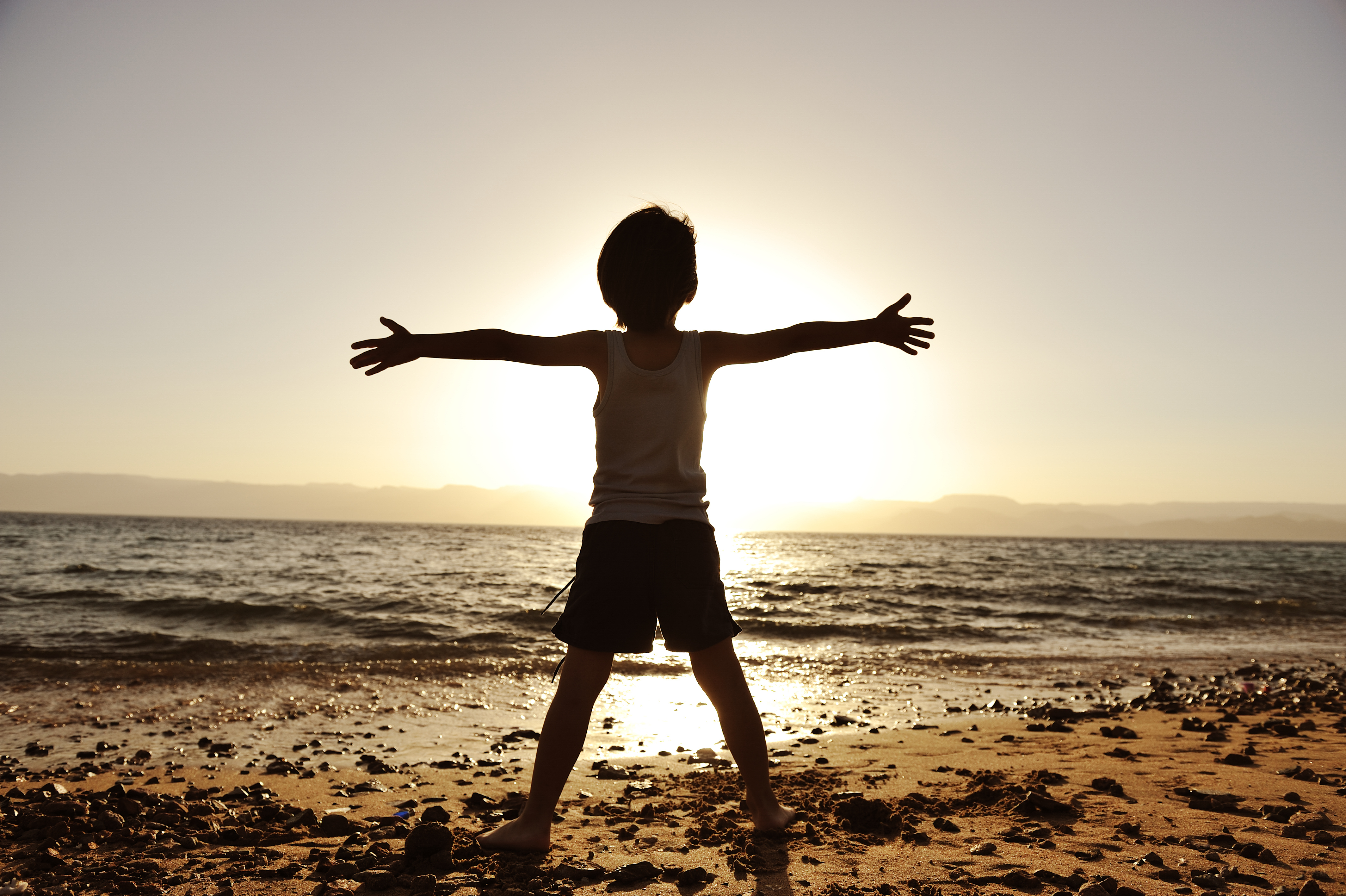 Silhouette of child on the beach, holding his hands up, hugging the su